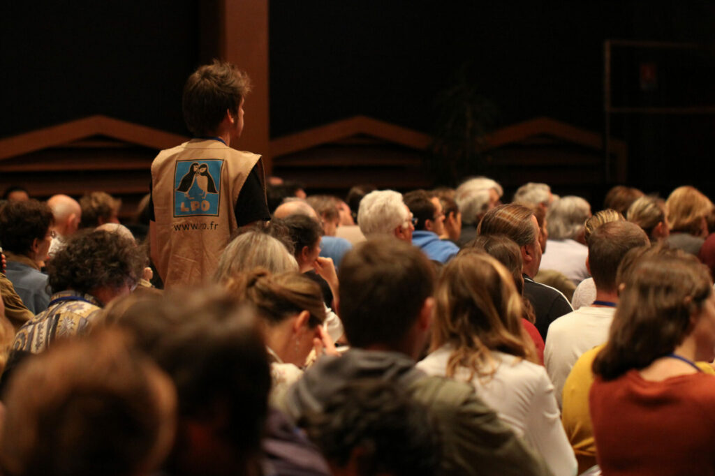 Une assemblée vue de dos ; une personne avec un gilet LPO est debout